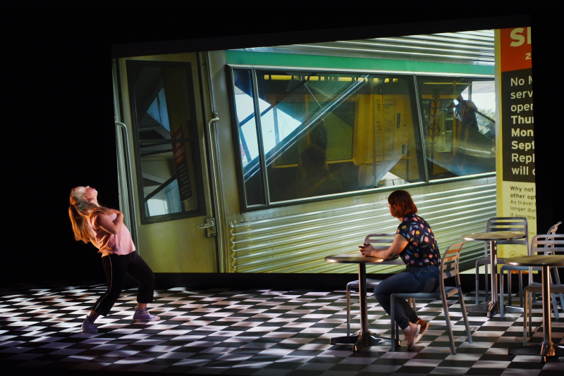 Julia sitting at a table set on a checkerboard floor with another actor standing