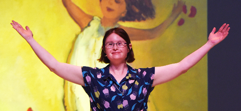 Julia Hales on stage with her arms in the air with an image of a little girl with her arms in the air behind her