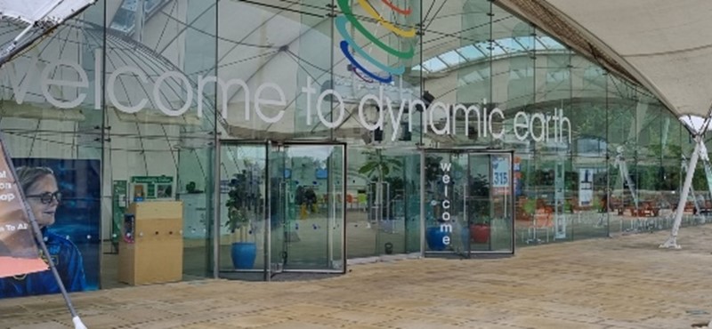 Photograph of the front entrance to Dynamic Earth featuring a canopy over glass front windows and doors and paved terrace