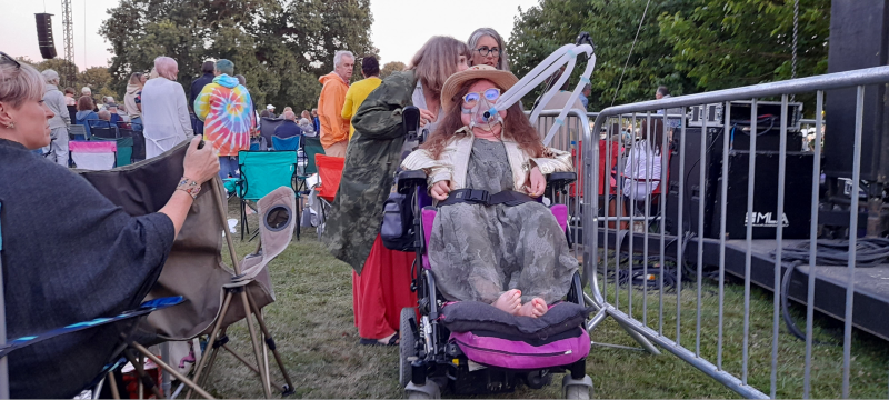 Karis in her power chair with people around her and a metal railing on grass