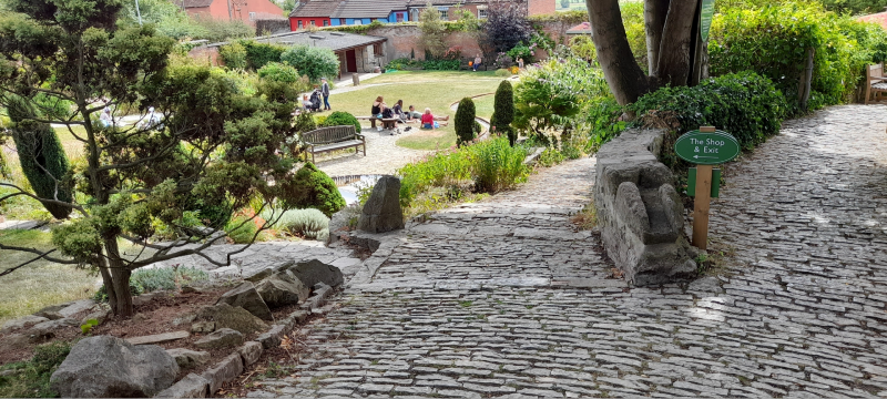 A cobbled path lined with trees and stones leading down a hill to grass lawns with people and benches with buildings in the background