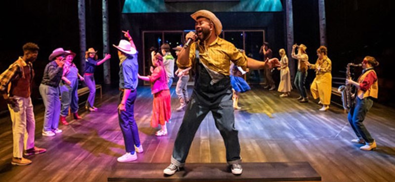 Richard is acting as the host of a line-dance and stands upon a bench, singing in to a microphone. Behind, the full company can be seen dancing in brightly-coloured costumes.