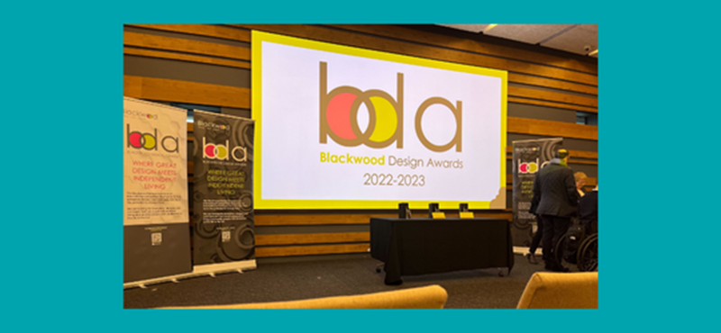 Teal background with a photograph of the Blackwood Design Awards branding on a screen on a stage with the tops of theatre style chairs just in view at the bottom front of photo