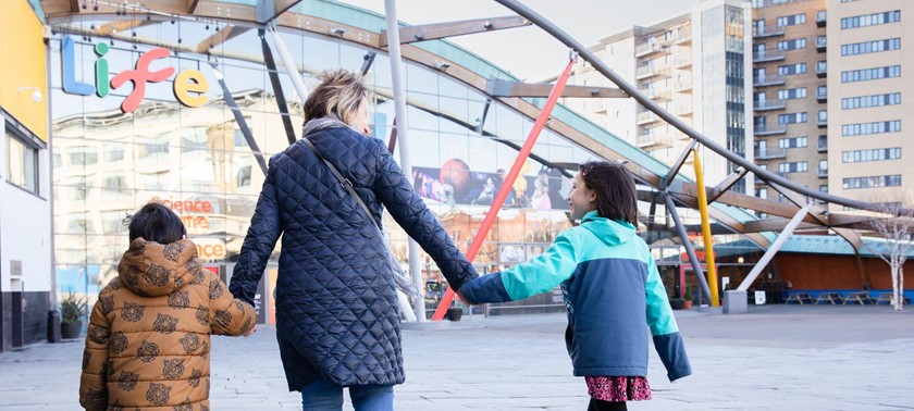 A photograph of a woman walking towards the Life Centre holding the hands of children on either side of her