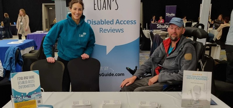 Euan's Guide staff member Christina and Euan's Guide Ambassador Jon smile to camera in front of a Euan's Guide pop up banner