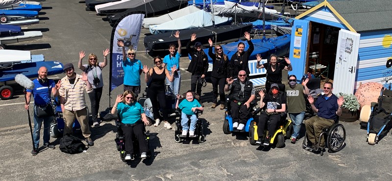 The group smiles and waves up to the camera which is positioned at a higher level with the Beach Wheelchairs hut and sailboats in the background