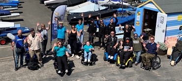 The group smiles and waves up to the camera which is positioned at a higher level with the Beach Wheelchairs hut and sailboats in the background