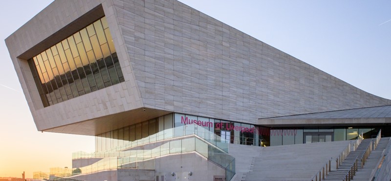 An external shot of Museum of Liverpool, a stone building with steps at the front and a ramp to the front left, and mirrored windows which reflect a sunset or sunrise