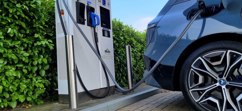An electric vehicle charging point with a car plugged in but the charging station is up on a kerb and has protective posts in the way of accessing the station