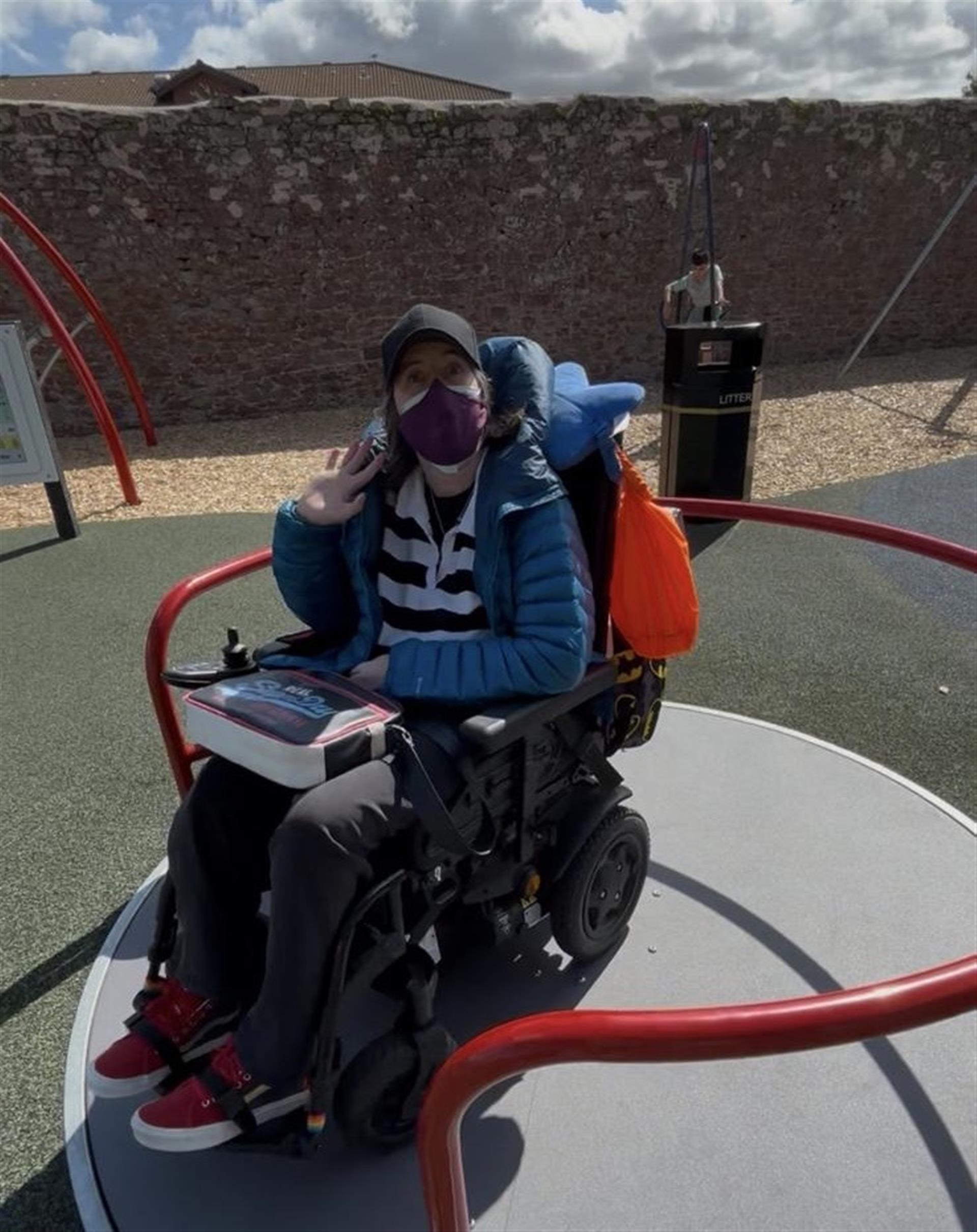 Euan's Guide Ambassador Karine sitting in her wheelchair, facing the camera with a mask on and waving  having fun on a wheelchair accessible roundabout