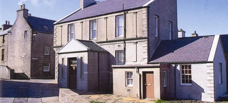 Photo of a large grey building next to a road with two entrance doors.