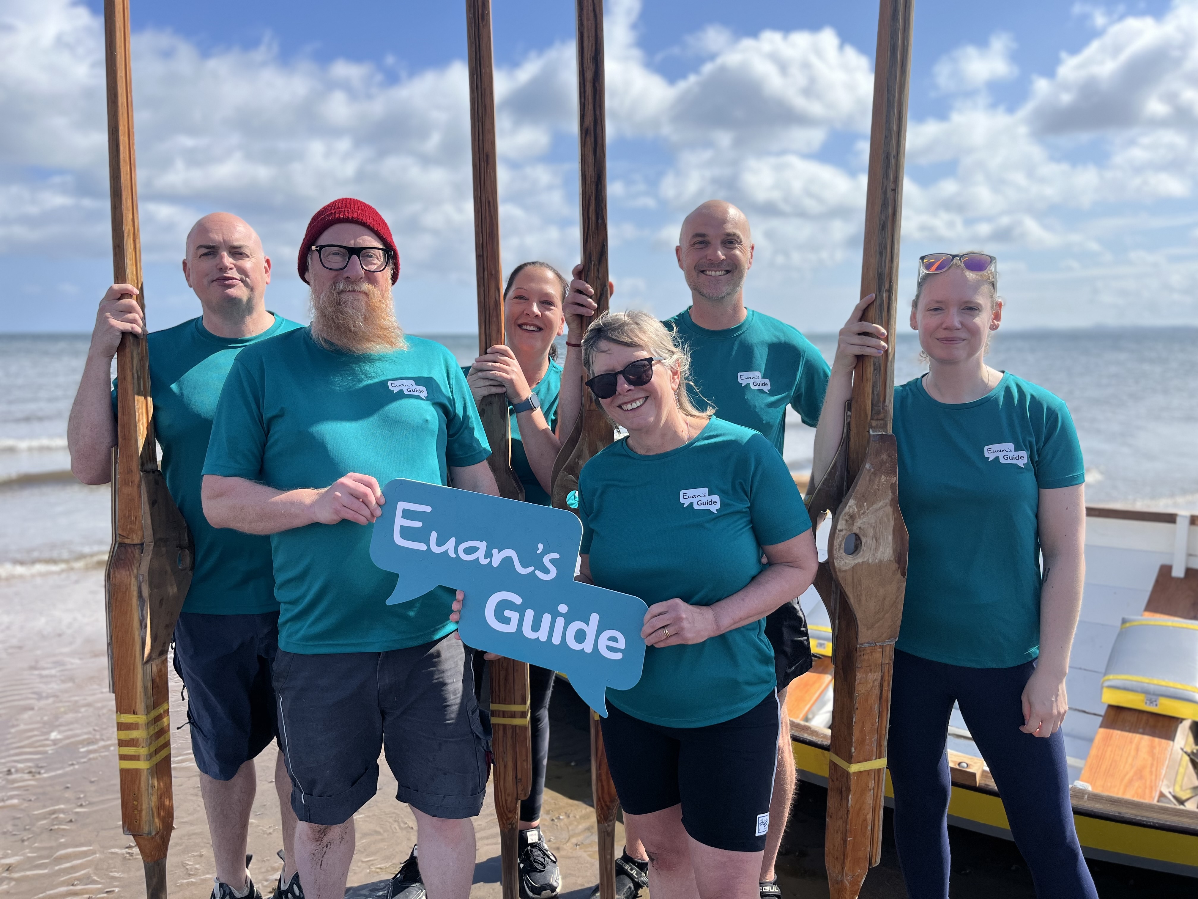 A photo of the rowing team up close holding a Euan's Guide logo prop and wearing teal Euan's Guide t shirts. They're holding their oars upright and behind them is the rowing boat, blue skies and the beach and sea