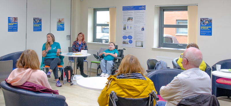 A group of people sit in a circle in an event space. The group consists of people sitting in chairs and people using wheelchairs and powerchairs