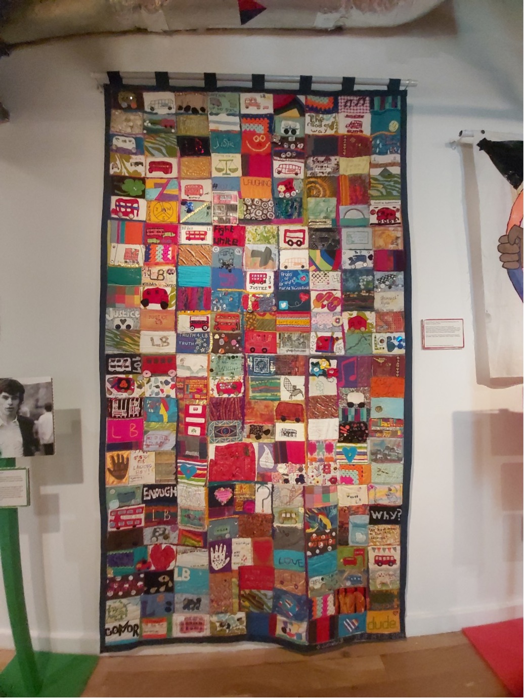 A photo of hanging piece of fabric art with lots of patchwork images sewn together against a white wall with other art just in shot