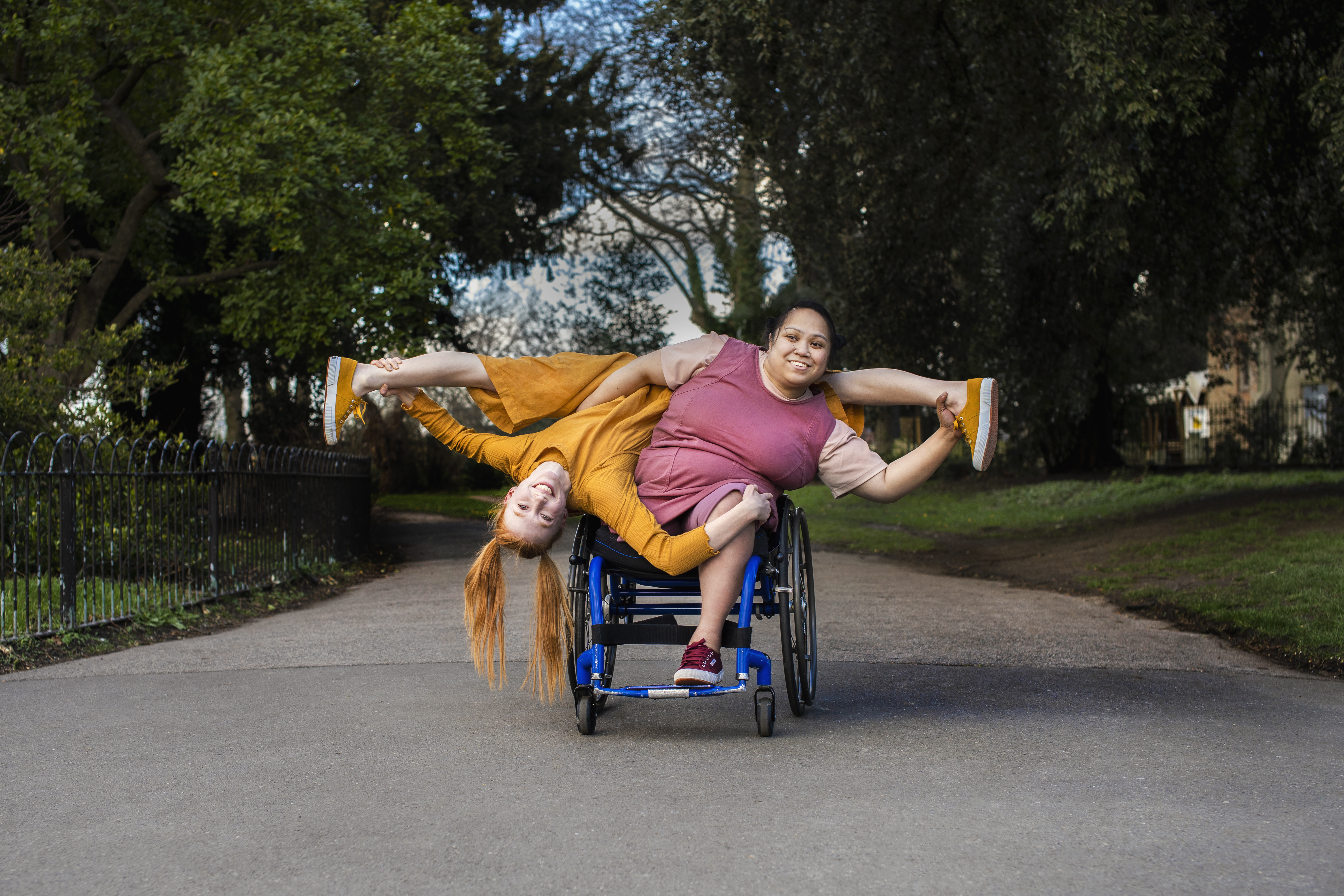 Two women create a pose, one woman uses a wheelchair and the other woman is held upside down alongside her
