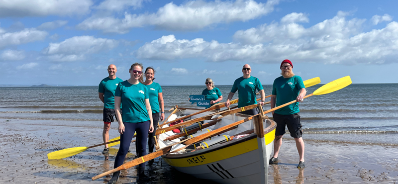 A photograph of the rowing team on the beach with their rowing boat and they're wearing teal Euan's Guide branded t shirts and holding a Euan's Guide logo prop