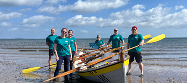 A photograph of the rowing team on the beach with their rowing boat and they're wearing teal Euan's Guide branded t shirts and holding a Euan's Guide logo prop