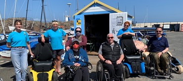 A photograph of Euan's Guide ambassadors with Beach Wheelchairs staff in front of the harbour with boats and boat sheds in the background