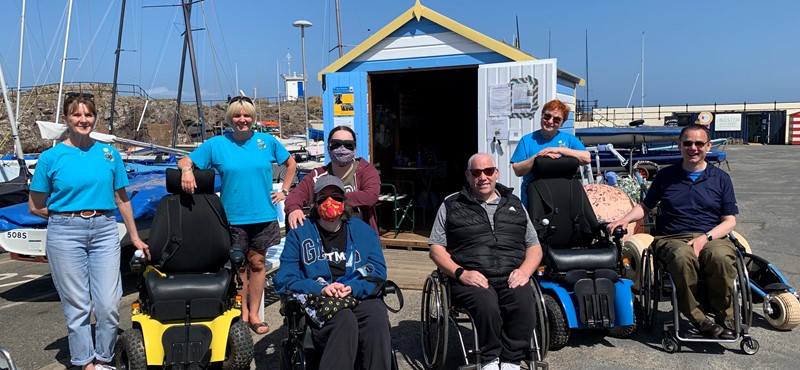 A photograph of Euan's Guide ambassadors with Beach Wheelchairs staff in front of the harbour with boats and boat sheds in the background
