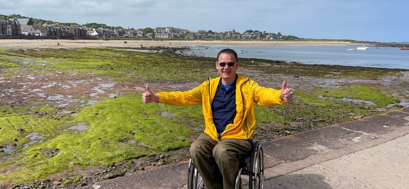 Paul Ralph, a wheelchair user, gives a big thumbs up as he smiles to camera from the pier at North Berwick. He wears a yellow jacket and the beach and sea is visible behind him.