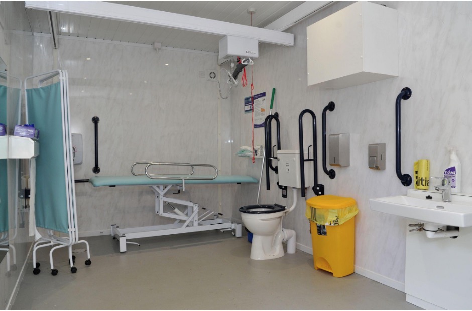 A photograph of the inside of a Changing Places Toilet with screen, changing bed, toilet, hoist, sink, yellow bin, grab rails and red emergency cord.
