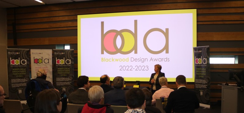 A photo of a stage with people sitting in front of it, with a digital display screen with the Blackwood logo on it