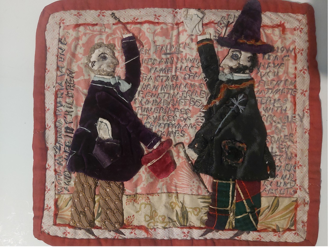A fabric sewn artwork with two men wearing coats and trousers one with a pointed hat