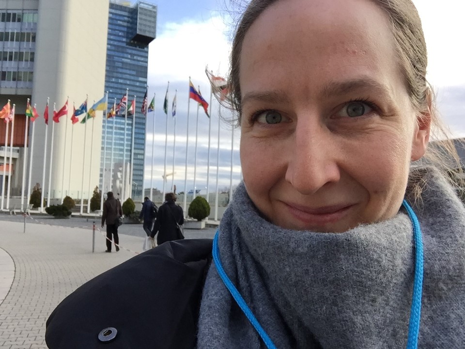 A selfie of Kat who has her hair tied back and a grey scarf on. There are flags in the background in front of a modern office block.