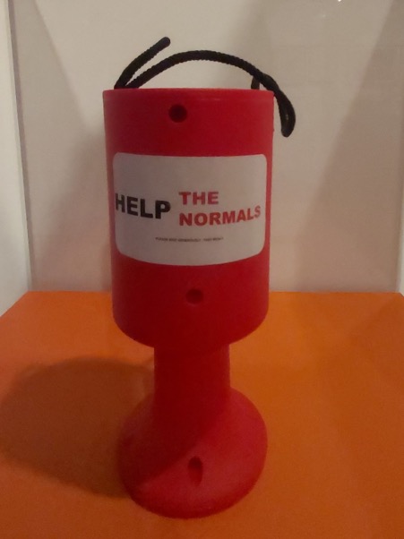 A photo of a red charity collection bucket with white label and black and red text 'help the normals'