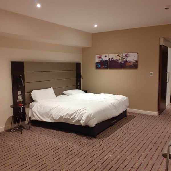 An accessable hotel room with a bed in the centre. On both sides there is a bed table with a light and a wall socket. On one side there is an emergency button to call for assistance. On the wall is some paintings with some flowers on it.