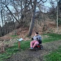 The image shows Pat, a white person with short brown hair wearing pink trousers and a gray jacket. They are in a power chair on a hillside looking at a sign in the shape of an open book. There are barren trees in the background.
