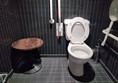 Accessible toilet cubicle, with a table obstructing the wheelchair transfer area and a red emergency cord much too short