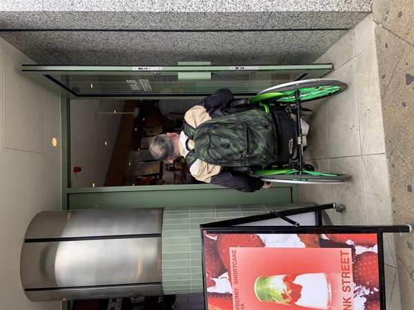 Image of a wheelchair user entering into the coffee shop through the door on the right. The door is not much wider than the wheelchair.
