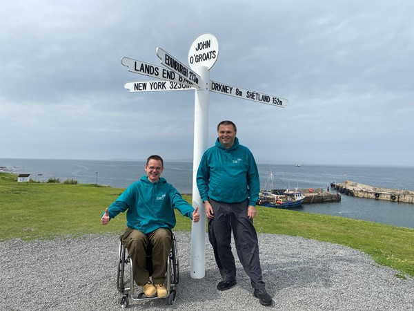 Paul and Ian from Euan's Guide standing alongside the John o' Groats signpost. They are both wearing teal coloured Euan's Guide hoodies.