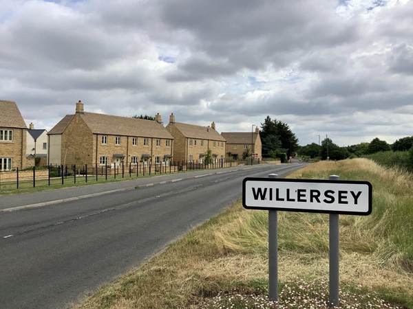 Picture of a sign saying "Willersey" in front of a road  and houses