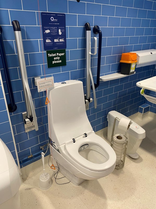 Photo of the toilet in the accessible changing places bathroom, it is a modern design and looks very new. It has built in Japanese style wash and dry bidet functions built in, and a remote control to operate them hung on the wall. A free standing toilet roll holder is in easy reach to the left of it and it can be moved around as needed. There are sanitary bins in easy reach of the toilet, the sink, and the washing table. There are grab bars on the walls either side of the toilet as well as fold-down grab bars as well, and a hoist above it. A sharps box sits on a shelf by the washing table