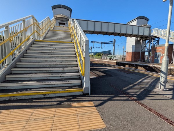 Image of of the foot of a stairway to the platform that crosses the tracks. There are rails on either side, tactile paving and yellow markings on the first and last step of each level