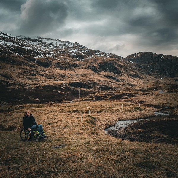 Helen is sitting in her manual wheelchair on grass, near a stream, with mountain views in the background. There's a little bit of snow on the hills and a moody, cloudy sky.