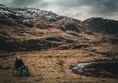 Helen is sitting in her manual wheelchair on grass, near a stream, with mountain views in the background. There's a little bit of snow on the hills and a moody, cloudy sky.