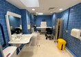 A changing places toilet, photo taken from the door, facing in. The walls are shiny blue tile, the floor is pale beige non slip. There is a bright yellow bin, paper towel dispensers, grab handles, adjustable height sink, soap dispenser, sanitary products bin, a folding screen a shower, a hoist, adjustable height washing table, toilet with built in bidet wash and dry functions with remote control to operate them, and a sharps box. There are three red cords around the large area, one of which has a Euan's Guide red cord card hung on it, and fold-away grab bars either side of the toilet. A powerchair is next to the toilet for scale