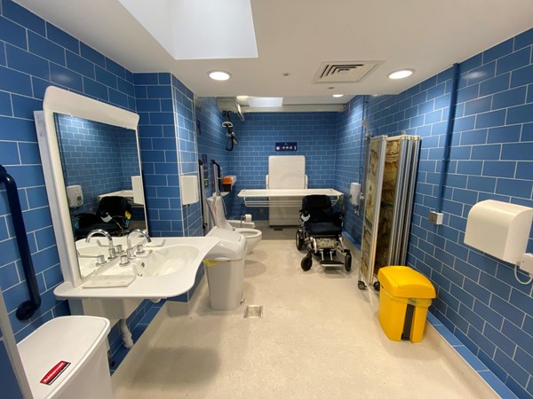 A changing places toilet, photo taken from the door, facing in. The walls are shiny blue tile, the floor is pale beige non slip. There is a bright yellow bin, paper towel dispensers, grab handles, adjustable height sink, soap dispenser, sanitary products bin, a folding screen a shower, a hoist, adjustable height washing table, toilet with built in bidet wash and dry functions with remote control to operate them, and a sharps box. There are three red cords around the large area, one of which has a Euan's Guide red cord card hung on it, and fold-away grab bars either side of the toilet. A powerchair is next to the toilet for scale