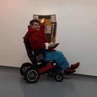 The image shows Pat, a white person with short brown hair wearing a red jacket and blue trousers. They are sitting in a powerchair and looking at the camera while reaching up to touch a quilt that is hung on the wall. The rest of the space is blank with white walls and grey flooring.