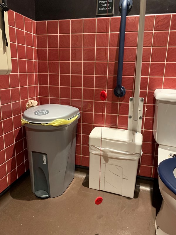 Image of the red cord hanging on the right side of the toilet. There is a small sign on the wall that reads "Please pull cord for assistance"