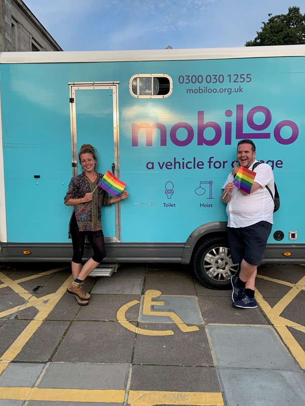 Mobiloo was in attendance - picture shows two people, a woman and a man, either side of the mobiloo sign on the mobiloo van. Each person is holding a pride rainbow flag.