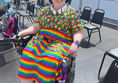 A wheelchair user wearing a rainbow dress, sunglasses and a hat is smiling, they are sitting on the wheelchair viewing platform with VIP areas behind them.