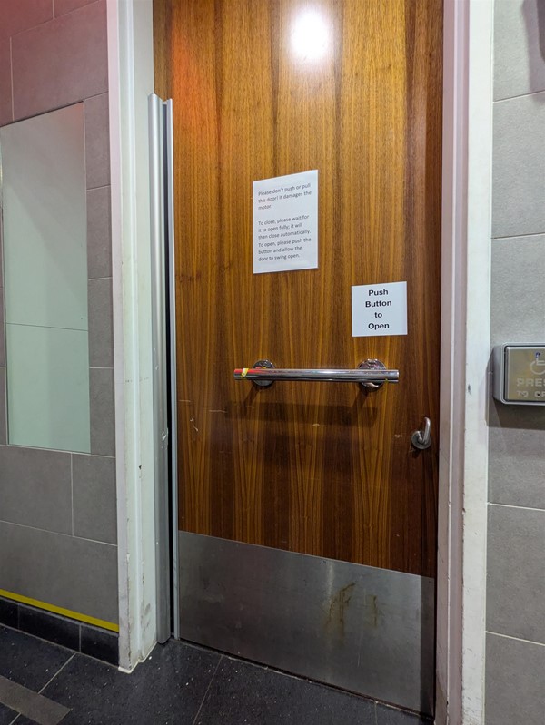 Image of accessible toilet door from the inside with horizontal grab rail, a button to open the door and a full-length mirror on the wall to the left