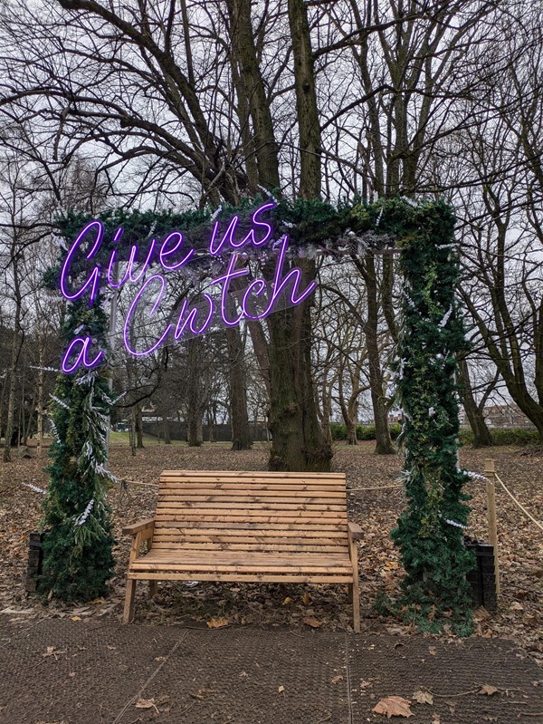 Image of a bench in the park, above which is a grass arch and the phrase 'Give us a Cwtch' in purple neon