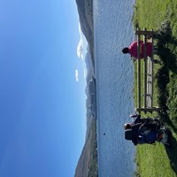 Photo of a male powered wheelchair user and a female sitting on a bench on grass with a walking aid in front of her. They are looking out towards a cruise ship on Lochbroom, the sun is shining and the sky and the loch are blue.