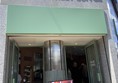 Image of the entrance from the outside. The facade is smooth grey stone and there are white letters over the door reading “BLANK STREET COFFEE”. There are two doors with mint-green frames next to each other with a metal and green-tiled pillar between them. There is a sign advertising a matcha drink. The doors are propped open and there are no steps.