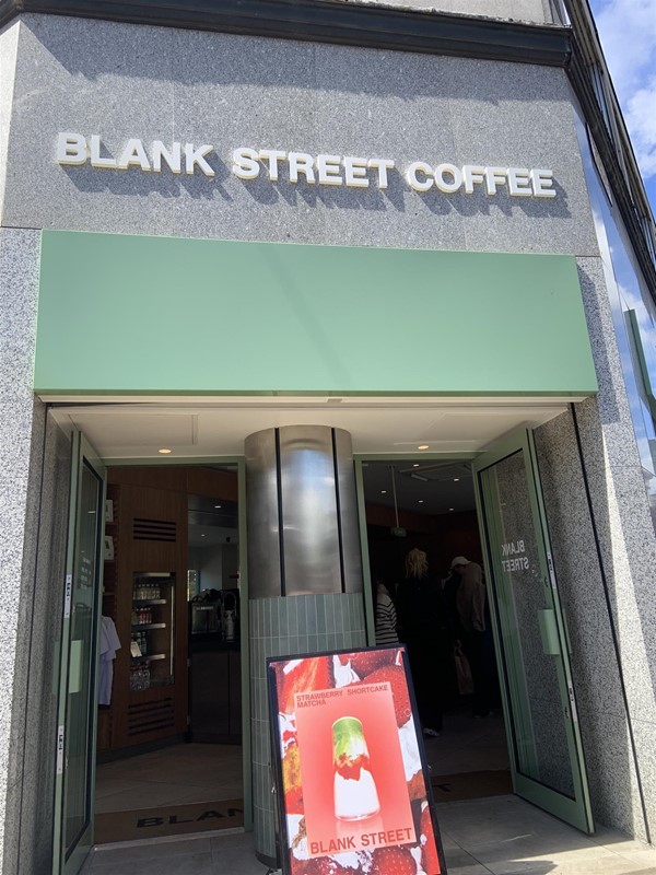 Image of the entrance from the outside. The facade is smooth grey stone and there are white letters over the door reading “BLANK STREET COFFEE”. There are two doors with mint-green frames next to each other with a metal and green-tiled pillar between them. There is a sign advertising a matcha drink. The doors are propped open and there are no steps.